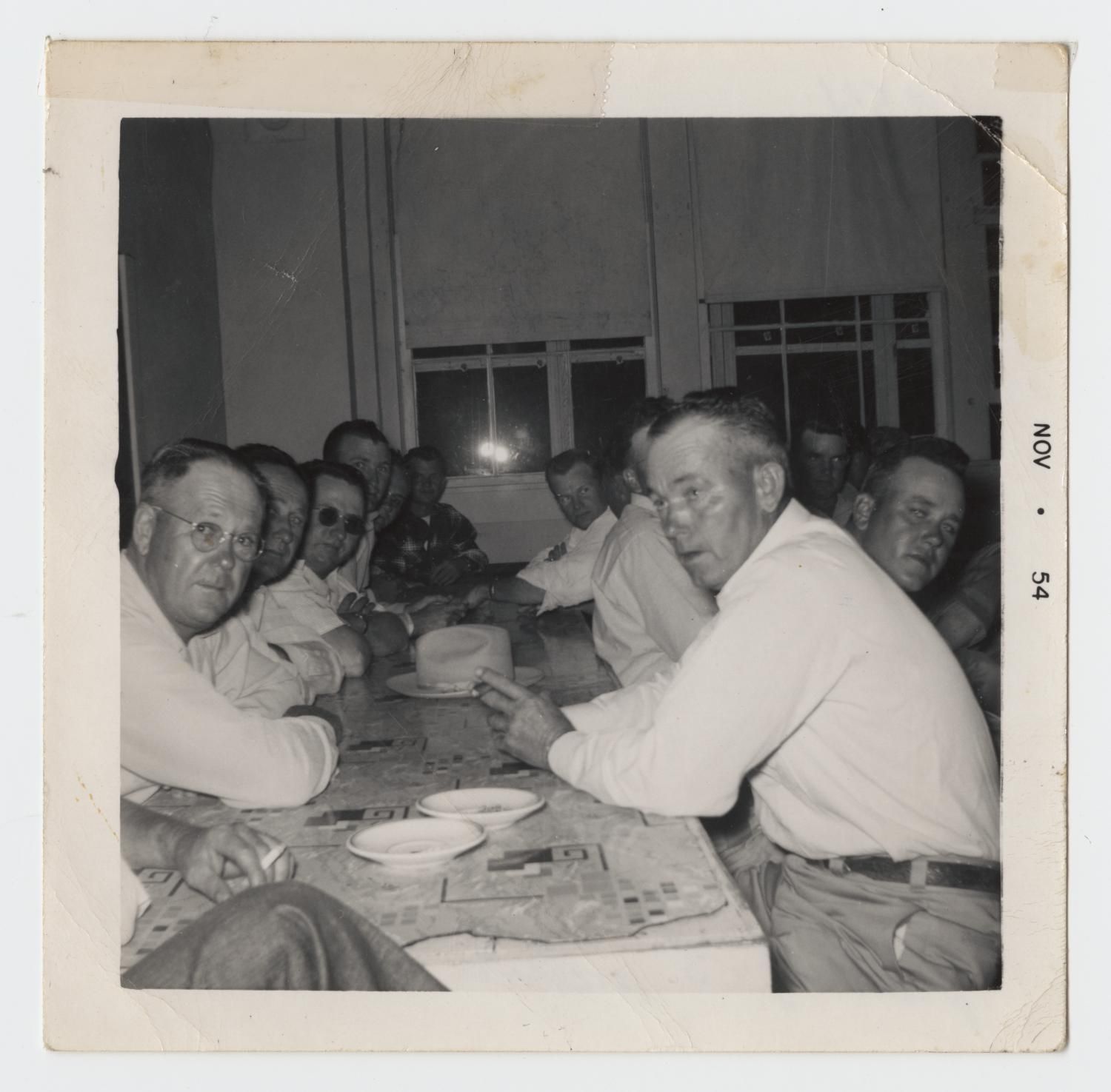 A long table of men in white shirts, several smoking, turn toward the camera during an evening meeting. The print is stamped "Nov 54."