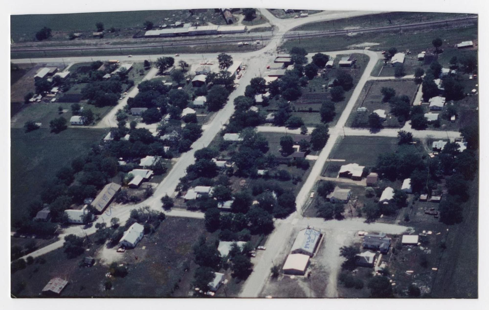An aerial color photograph of a small town grid: tree-lined streets, scattered houses and low buildings, and a railroad line running across the top.